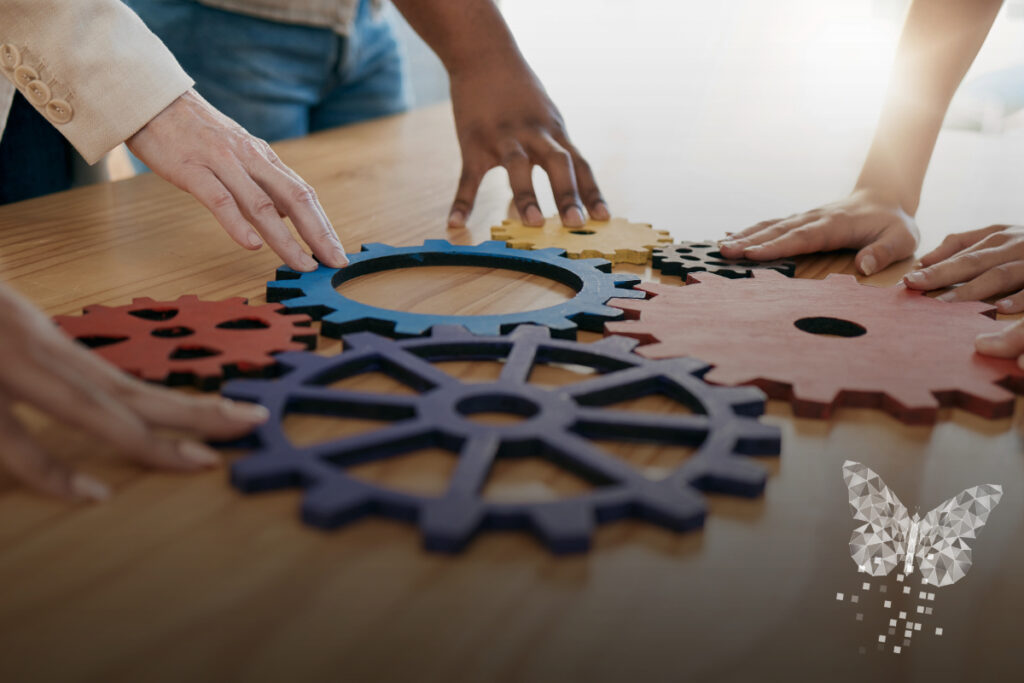 Several hands of diverse individuals collaborate to connect large, colorful wooden gears on a table, symbolizing teamwork and cooperation. A polygonal butterfly graphic appears in the lower right corner, adding a creative or transformational element to the composition.