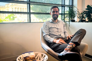 In a modern office with potted plants and city views, a cheerful man sits relaxed in a gray chair, crossing his legs to reveal bold, colorful socks. A “Health Policy” pamphlet lies on a small round table in front of him.
