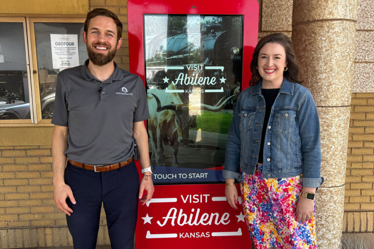 A man and woman stand smiling in front of a red interactive display kiosk that reads “Visit Abilene Kansas,” with an image of a train and longhorn cattle in the background. The man wears a gray polo shirt and navy pants, and the woman wears a denim jacket over a colorful floral skirt.