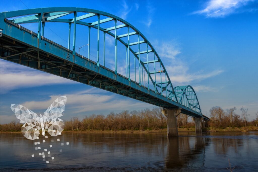 A long, arched blue bridge crosses a Kansas river lined with leafless trees. In the foreground, a semi-transparent, polygonal butterfly design hovers above the water.